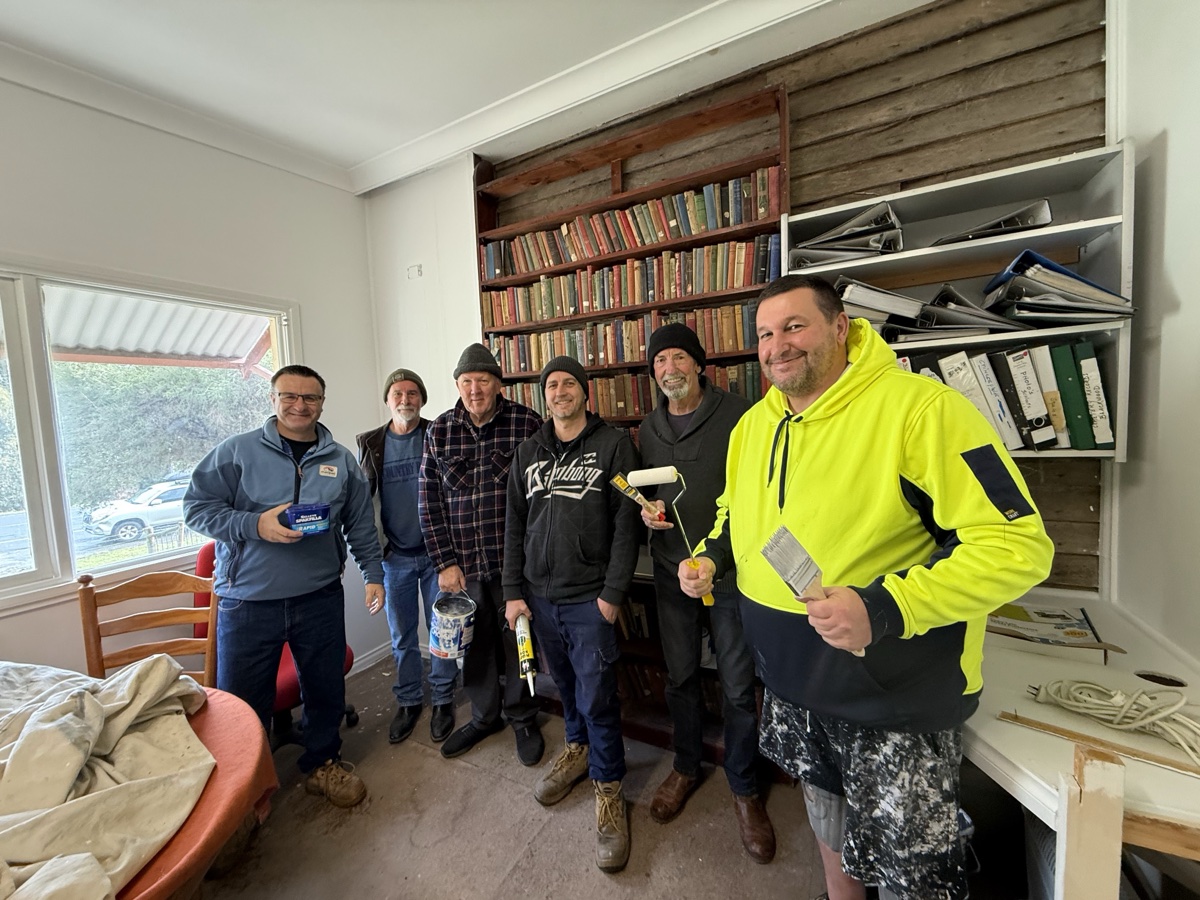 Seven Blackwood & District Historical Society volunteers holding paint brushes, rollers and tins in the freshly painted meeting room, standing in front of the timber bookshelves and archive files